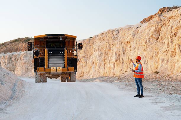 Foreman wearing an orange vest and hardhat directing a dumper truck in a quarry using a tablet